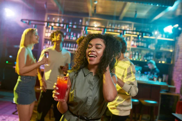 Atractiva mujer joven de raza mixta sonriendo a la cámara mientras posa con un cóctel en la mano y amigos charlando, tomando bebidas en el mostrador del bar en el fondo — Foto de Stock