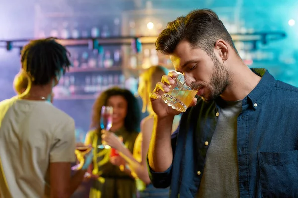 Joven guapo bebiendo un cóctel mientras sus amigos charlan, tomando bebidas en el mostrador del bar en el fondo — Foto de Stock