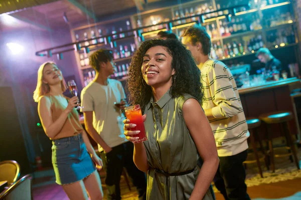 Disfruta de la vida nocturna. Atractiva joven mestiza sonriendo a la cámara mientras posa con un cóctel en la mano. Amigos charlando, tomando bebidas en el mostrador del bar en el fondo — Foto de Stock