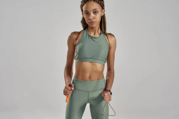 Joven mujer de carrera mixta deportiva en ropa deportiva sosteniendo saltar la cuerda y mirando a la cámara mientras está de pie sobre un fondo gris en el estudio — Foto de Stock