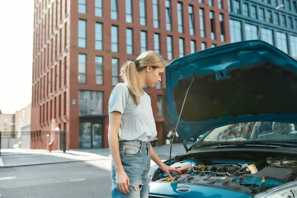 Aantrekkelijke jonge vrouw onderzoeken, kijken naar haar kapotte auto met open motorkap terwijl staan op de straat — Stockfoto