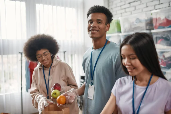 Retrato de un joven voluntario sonriendo a la cámara mientras empaca la donación de alimentos y bebidas en bolsas y cajas de papel, equipo pequeño que trabaja en la fundación benéfica — Foto de Stock