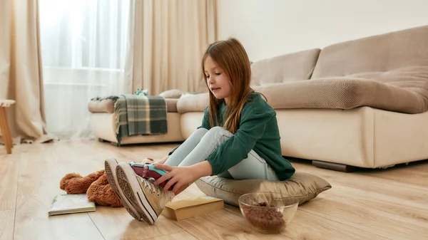 A little cute girl hutting a gamepad on her feet smiling sitting on a floor of a big guestroom — Stock Photo, Image