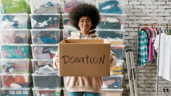 寄付箱を持っているアフリカ系アメリカ人の女の子とカメラを見て、服でいっぱいの箱の前にポーズをとって、慈善団体のために働く若いボランティア — ストック写真