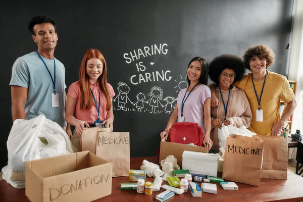 Grupo de diversos jóvenes voluntarios sonriendo a la cámara mientras empaquetan pastillas, donación de medicamentos, compartir es cuidar la inscripción en el fondo, equipo pequeño que trabaja en la fundación caritativa — Foto de Stock