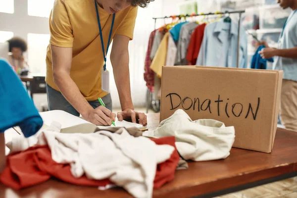 Beskuren bild av unga manliga volontär att göra anteckningar medan du bockar kläder i donationslåda för behövande människor, Selektivt fokus på hand — Stockfoto