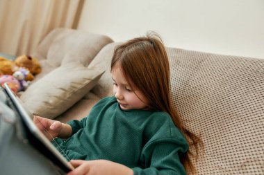 Küçük bir kız tabletinde video oyunları oynamayı seviyor. Kanepeye uzanmış, ekrana bakıyor.