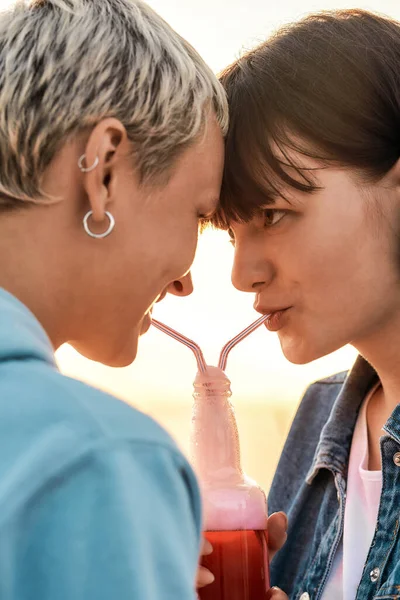 Zbliżenie młodej lesbijskiej pary patrzącej na siebie, pijącej z jednej szklanej butelki ze słomką, Dwie kobiety cieszące się zimnym napojem w letni dzień na świeżym powietrzu — Zdjęcie stockowe