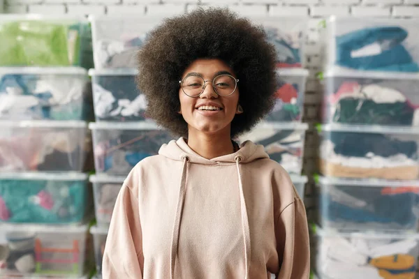 Ritratto di felice ragazza afroamericana che sorride alla telecamera mentre posa davanti a scatole piene di vestiti, Giovane volontaria che lavora per una carità, donando abbigliamento alle persone bisognose — Foto Stock