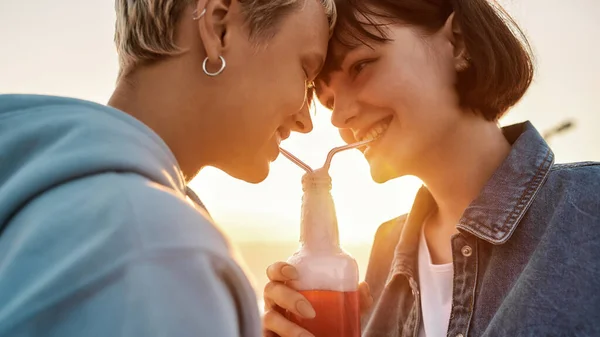 Cropped shot of happy young lesbian couple drinking from one glass bottle with the straw, Two women enjoying cold beverage on a summer day outdoors — Stock Photo, Image