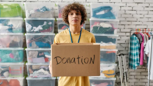 寄付箱を持っている白人男性とカメラを見て,ラックや服でいっぱいの箱の前にポーズ,慈善のために働く若いボランティア — ストック写真