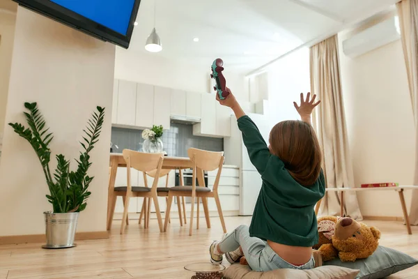 Una niña pequeña sentada en el suelo frente a un televisor vitoreando después de ganar un videojuego sosteniendo un mando en una gran habitación luminosa —  Fotos de Stock