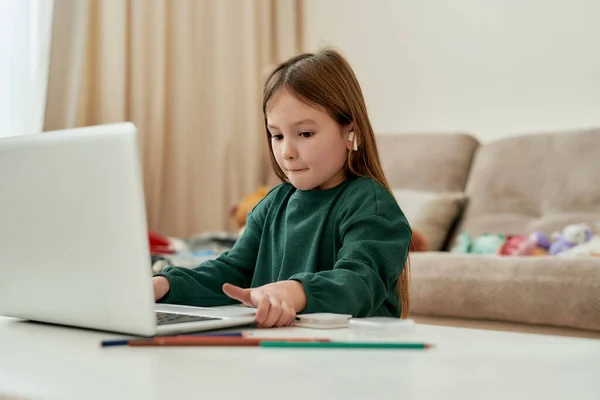 Uma garota bonita decidiu fazer sua tarefa de educação à distância e mover um laptop em uma mesa enquanto se senta sozinha em um grande quarto brilhante — Fotografia de Stock