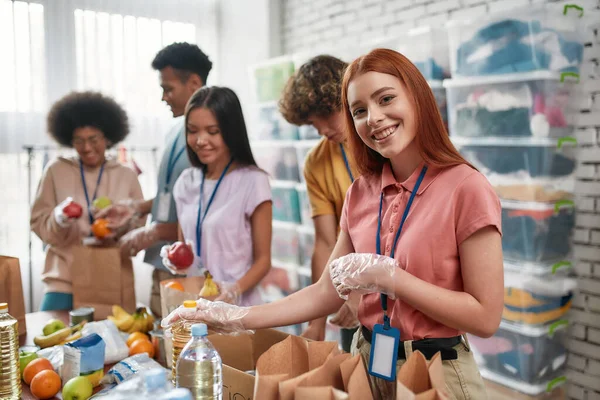 Jonge vrouwelijke vrijwilliger glimlachend op camera tijdens het verpakken van voedsel en dranken donatie in papieren zakken en doos, Kleine groep mensen die werken in liefdadigheidsstichting — Stockfoto