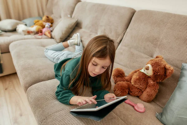 Uma menina fofa pequena que joga jogos em seu tablet que jaz em seu estômago em um sofá com seus gumshoes sobre e um brinquedo fofo ao redor — Fotografia de Stock