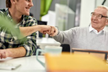 Yaşlı bir adam, kıdemli bir stajyer genç meslektaşına gülümseyip yumruk tokuştururken yeni çalışana ofiste yardım ederken arkadaşça davranan bir erkek.