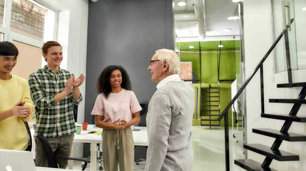 Hombre anciano, pasante senior mirando complacido mientras sus colegas lo elogian en el primer día en el trabajo, trabajadores amigables aplaudiendo, animando a un nuevo empleado en la oficina — Foto de Stock