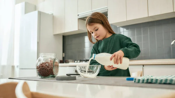 Ein süßes kleines Mädchen gießt Milch in eine Schüssel, während es in einer großen, gut beleuchteten Küche steht — Stockfoto