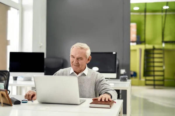 Portrait of cheerful aged man, senior intern looking focused at the screen while using laptop, sitting at desk, working in modern office — Stock Photo, Image