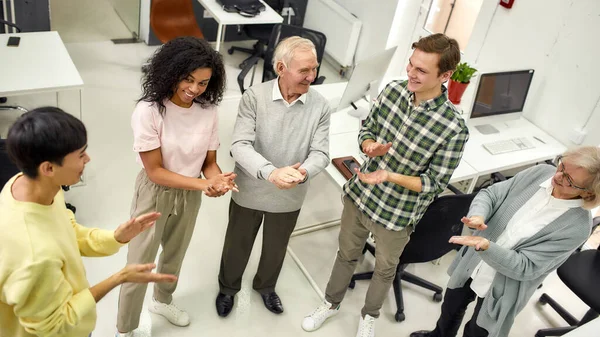 Gruppe unterschiedlicher Mitarbeiter applaudiert, während sie zusammen im Büro stehen, Alte Frau und Mann, Senior-Praktikanten beim ersten Arbeitstag — Stockfoto