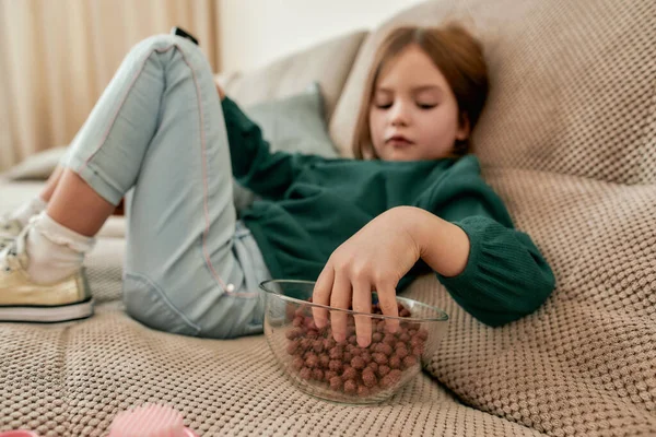 Una niña pequeña sentada pierna a pierna en un sofá comiendo bolas de cereal de un tazón de cristal en una habitación luminosa —  Fotos de Stock