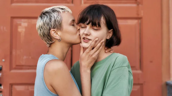 Pretty woman with short hair kissing her girlfriend outdoors. Young female couple standing in front of the door. LGBT, Sexual freedom concept — Stock Photo, Image