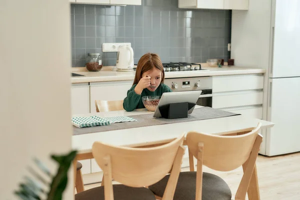 Uma menina está assistindo um tablet atentamente enquanto está sentada sozinha em uma mesa e comendo seu café da manhã rápido com uma colher em uma cozinha grande brilhante em casa — Fotografia de Stock