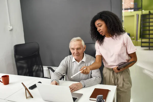 Aged man, senior intern looking at laptop while listening to his young colleague, Friendly female worker training or teaching new employee, preparing for first day at work — Stock Photo, Image