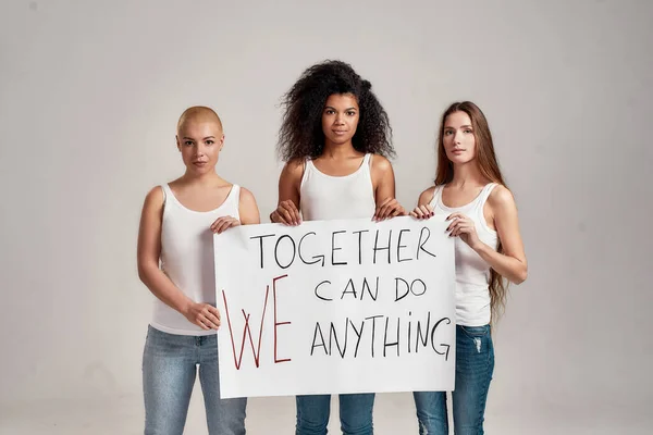 Tres mujeres seguras y diversas en camisa blanca y vaqueros casuales sosteniendo, de pie con una pancarta en sus manos aisladas sobre fondo gris — Foto de Stock