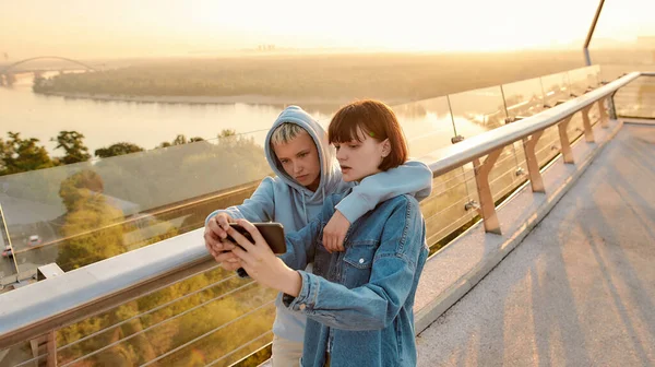 Pareja de lesbianas de pie en el puente, tomando una foto selfie mientras observan el amanecer juntos —  Fotos de Stock