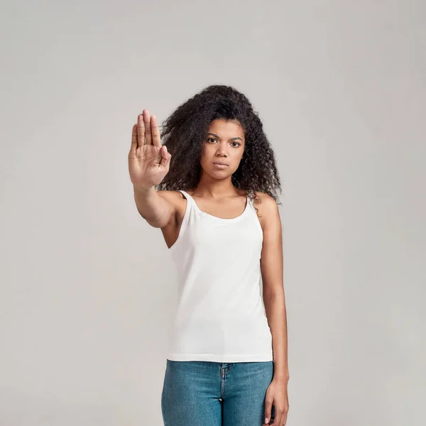 Retrato de jovem afro-americana com cabelo encaracolado vestindo camisa branca com sério olhar confiante ao mostrar, fazendo parar gesto isolado sobre fundo cinza — Fotografia de Stock