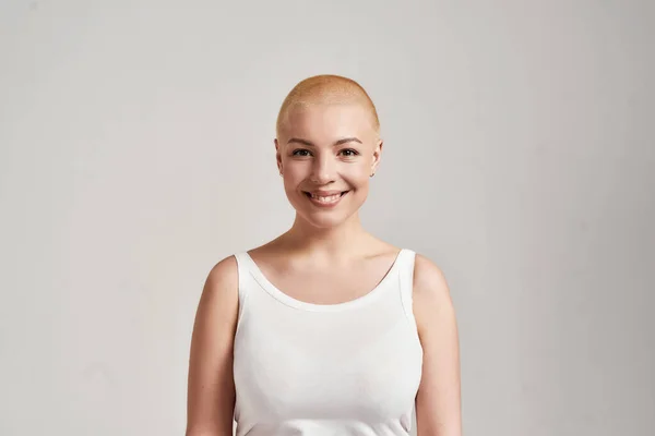 Retrato de uma bela jovem caucasiana com a cabeça raspada vestindo camisa branca, sorrindo para a câmera enquanto posando isolado sobre fundo cinza — Fotografia de Stock