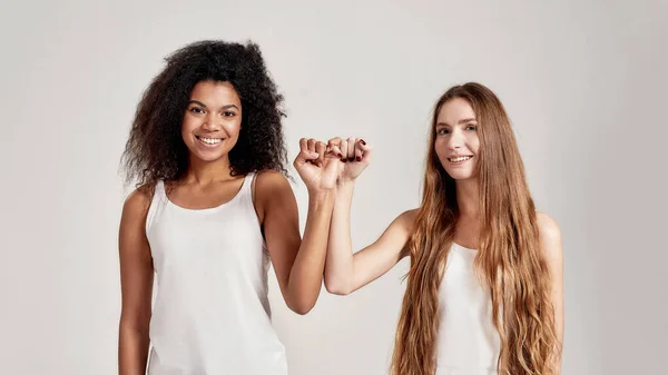 Portrait of two young diverse women, best friends smiling at camera while making a pinkie promise sign isolated over grey background — Stock Photo, Image
