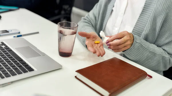 Close up of hands of aged woman, senior intern holding, taking pills out of bottle while using laptop, sitting at desk, working in modern office — Stock Photo, Image