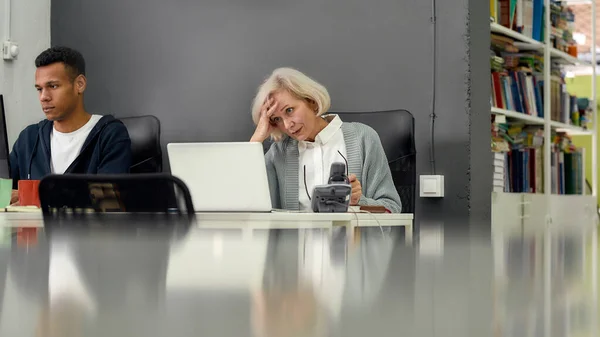 Mujer anciana, pasante senior con aspecto incierto, confundido en la pantalla mientras usa el ordenador portátil, sentado en el escritorio, trabajando en la oficina moderna con otro joven empleado —  Fotos de Stock