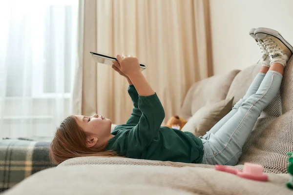 Маленька мила дівчинка грає в ігри на планшеті, лежачи на дивані з її ногами вгору і камерою на — стокове фото