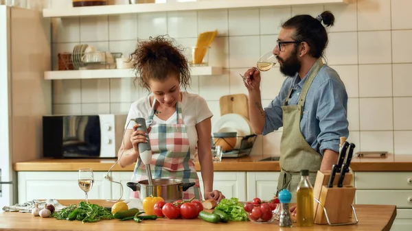 Uomo italiano, cuoco cuoco bere vino bianco mentre la donna utilizzando frullatore a mano. Giovane coppia che prepara un pasto insieme in cucina. Cucina casalinga, Cucina italiana — Foto Stock