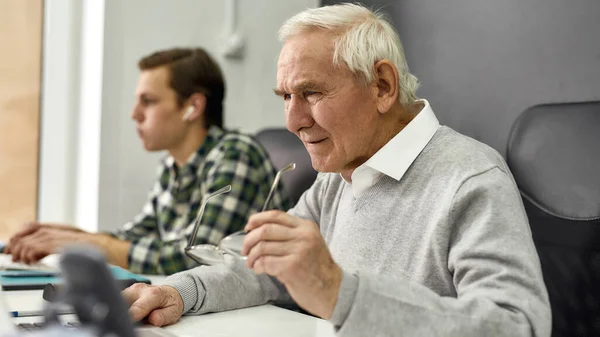 Close up of aged man, senior intern looking focused while using laptop, sitting at desk, working in modern office with young colleague — Stock Photo, Image