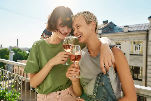 Две веселые женщины держат бокал вина, обнимаются во время вечеринки, празднуют годовщину на балконе. Молодая лесбийская пара проводит свой день вместе — стоковое фото