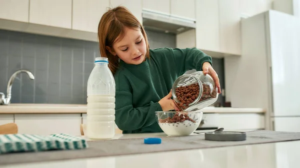 Ein kleines nettes Mädchen gießt in einer großen, hellen Küche Müslibällchen in eine Schüssel mit Milch — Stockfoto