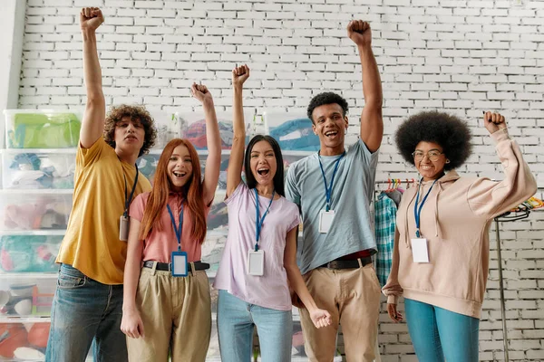 Giovani diversi gruppi di volontari alzarono le braccia mentre posavano davanti a scatole piene di vestiti, squadra felice che lavora per una carità, donando abbigliamento alle persone bisognose — Foto Stock