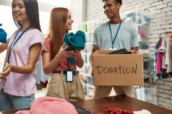 Felices jóvenes voluntarios clasificando, empacando ropa en cajas de cartón, Equipo diverso trabajando juntos en el proyecto de donación en interiores — Foto de Stock