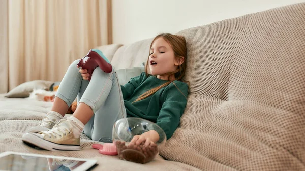 Una pequeña chica linda comiendo bolas de cereal mientras juega videojuegos sosteniendo un mando sentado en un sofá en una habitación grande —  Fotos de Stock