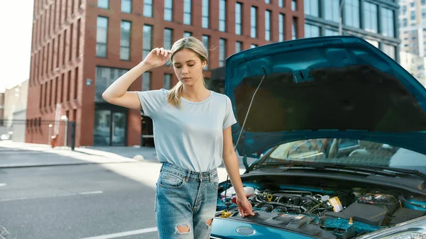 Atrakcyjna młoda kobieta pozowała stojąc w pobliżu zepsutego samochodu z otwartym kapturem na ulicy miejskiej — Zdjęcie stockowe