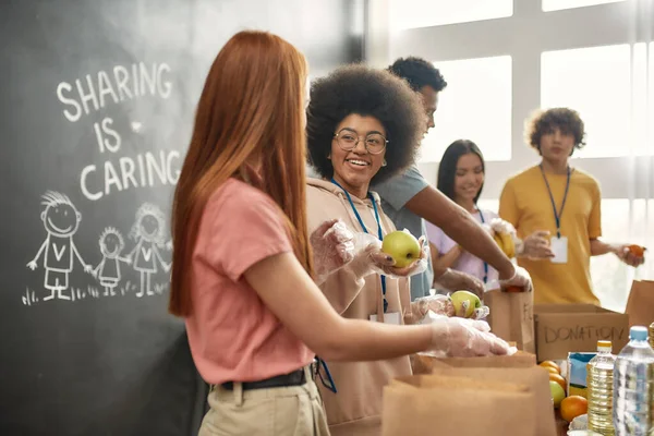 Glada unga volontärer i handskar samla in, sortera mat för behövande människor, Diverse team arbetar tillsammans med donation projekt i välgörenhet organisationskontor — Stockfoto