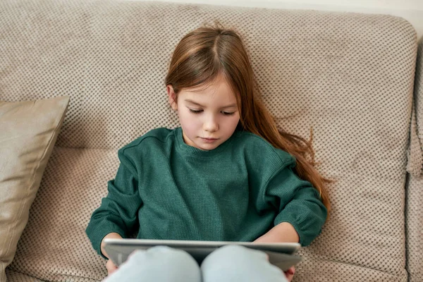 Uma menina fofa pequena envolvida em um processo de jogar jogos em um tablet enquanto sentado em um sofá acolhedor — Fotografia de Stock