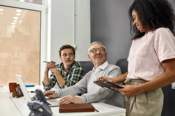 Homem idoso, estagiário sênior sorrindo para seu jovem colega enquanto usa laptop, Trabalhadores amigáveis mentoria e treinamento de novo funcionário, monitorando seu progresso no trabalho — Fotografia de Stock
