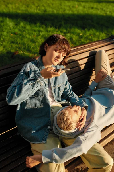 Young woman taking picture of her girlfriend while relaxing on the bench in the city park on a sunny day. Lesbian couple spending time together outdoors
