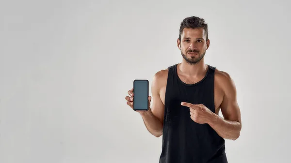 Schöner, athletischer Mann zeigt auf sein Smartphone mit leerem Bildschirm und schaut in die Kamera, während er isoliert im Studio vor grauem Hintergrund steht und Fitness-App nutzt — Stockfoto
