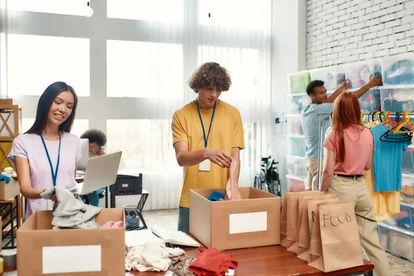 Mladí dobrovolníci skupinové třídění, balení potravin a oblečení v kartonových krabicích, Diverse tým pracuje na projektu darování uvnitř — Stock fotografie